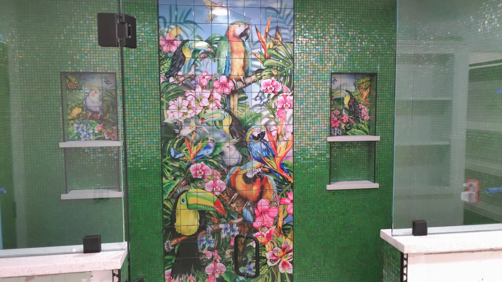 Green mosaic tile shower with tropical bird and orchid mural, white shelves, and coordinating tile panels